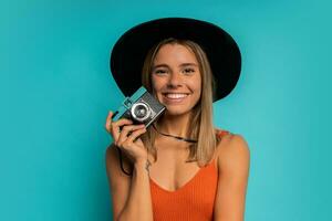 mooi lachend vrouw in zwart hoed en oranje top Holding wijnoogst camera en poseren over- turkoois achtergrond in studio. foto