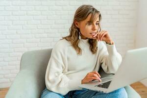 binnen- levensstijl foto van mooi vrouw met blond golvend haren zittend Aan sofa Bij huis werken Aan laptop computer. vervelend knus trui.