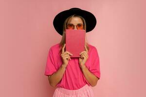 schattig blond vrouw in zwart hoed en roze voorjaar kleding Holding boek en poseren in studio. foto