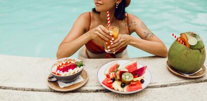 zelfverzekerd brunette bruinen vrouw in rood kat ogen zonnebril ontspannende in zwembad met bord van exotisch fruit gedurende tropisch vakantie. elegant tatoeëren. foto