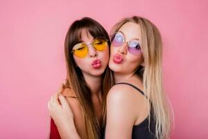 paar van mooi meisjes, het beste vrienden poseren Aan roze achtergrond. twee Dames in kleurrijk zonnebril sturen kus. foto