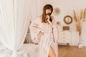 sensueel brunette vrouw poseren in beige zijde kimono in elegant licht slaapkamer in boh stijl. foto