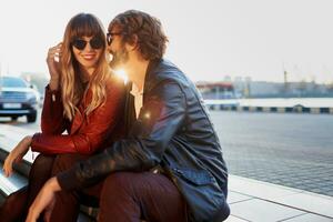 buitenshuis mode beeld van elegant paar in gewoontjes kleding, leer jasje en zonnebril zittend Aan de modern straat. knap Mens met baard met zijn vriendin uitgeven romantisch tijd samen. foto