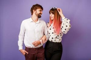 schattig paar knuffelen en op zoek Aan elk ander. knap Mens met baard en zijn mooi vrouw met roze haren in elegant retro kleding poseren Aan Purper achtergrond in studio. foto