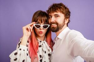 mooi vrouw met openhartig glimlach en roze haren poseren met haar vriendje met baard. hipster paar in liefde Aan Purper achtergrond. wijnoogst zonnebril. foto