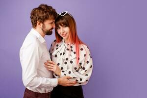 schattig paar knuffelen en op zoek Aan elk ander. knap Mens met baard en zijn mooi vrouw met roze haren in elegant retro kleding poseren Aan Purper achtergrond in studio. foto