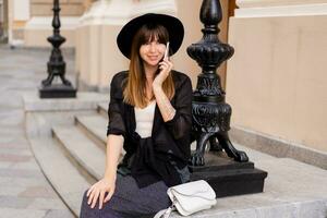 prachtig bruin vrouw in elegant herfst kleding en terug hoed pratend door mobiel telefoon Aan de straat in oud Europese stad. foto