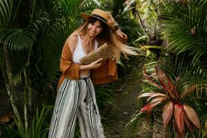 mode portret van brunette vrouw in rietje hoed poseren Aan tropisch palm bladeren achtergrond in Bali. vervelend elegant Boheems accessoires. foto