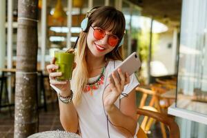 elegant vrouw in roze bril genieten van groen gezond smoothie , luisteren muziek- door oortelefoons, Holding mobiel telefoon. verrassing gezicht. modieus accessoires. tropisch humeur. foto