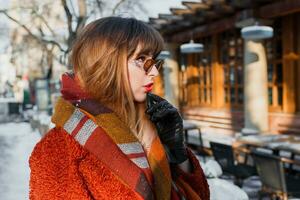 dichtbij omhoog winter portret van elegant brunette vrouw in retro bril poseren buitenshuis in oud Europese stad. staand in de buurt cafe. vervelend bruin jas, wol sjaal en leer handschoenen. foto