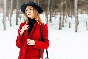 winter mode portret van blond leerling vrouw in hoed en rood jas wandelen in sneeuw park met tas. verkoudheid seizoen. foto