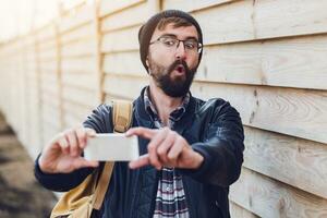 verrast gezicht. hipster Mens maken selfie Aan smartphone, gek emoties . poseren Aan hout muur achtergrond. vervelend elegant zwart hoed, leer jasje en rugzak. foto