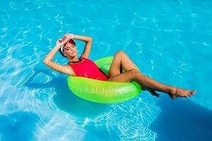 sexy gebruind vrouw in rood zwempak hebben pret en genieten van zomer in verbazingwekkend groot zwemmen zwembad. jong mooi meisje zwemmen Aan opblaasbaar ring. elegant transparant kap. strand feest. foto