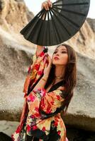 mode foto van elegant Aziatisch vrouw in zijde kimono Holding ventilator en poseren over- rotsen Aan de strand.