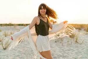 zomer humeur. speels brunette vrouw dansen in zonlicht Aan de strand. boho stijl. modieus halsketting en oorbellen. foto