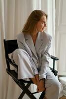binnen- mode foto van elegant blond vrouw in gewoontjes pak zittend Aan stoel. golvend haren, licht modern interieur.