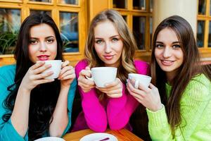 dichtbij omhoog levensstijl portret van drie mooi jong Dames zittend in cafe en genieten van heet tee . vervelend helder neon geel , roze en blauw elegant trui .vakantie, voedsel en toerisme concept . foto