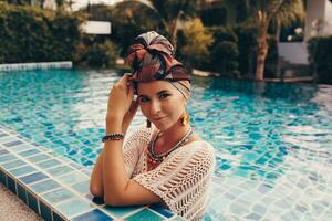 vrouw met helder maken omhoog in Boheems kleding, tulband, etnisch oorbellen, ketting en armbanden poseren in zwemmen zwembad. foto