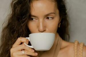 dichtbij omhoog portret van mooi brunette vrouw met perfect golvend haren genieten van kop van koffie in modern cafe. foto