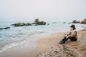 vrouw toerist in hoed zittend op het strand kijken naar zee foto