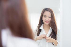jonge aziatische vrouw die met gezicht en glimlach onderzoekt die op spiegel kijken.