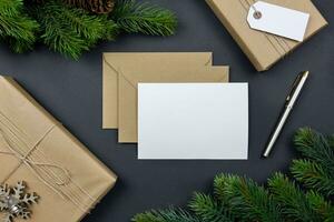 Kerstmis groet kaart en envelop mockup met handgemaakt geschenk doos, pijnboom kegels en Spar boom takken Aan donker achtergrond. feestelijk gestileerd samenstelling. top visie. foto