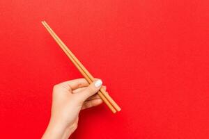 Bijsnijden beeld van vrouw hand- Holding eetstokjes Aan rood achtergrond. Japans voedsel concept met kopiëren ruimte foto