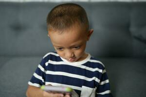 Aziatisch jongen spelen met cel telefoon Aan sofa binnen huis foto