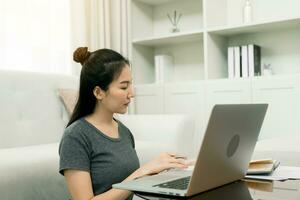 jonge aziatische vrouw zit in de woonkamer met behulp van een rekenmachine om gezinsinkomen en -uitgaven te berekenen en schrijft het thuis op in zijn notitieboekje of laptop. foto