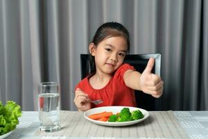 schattig weinig meisje tonen duim tonen aan het eten gezond groenten. foto