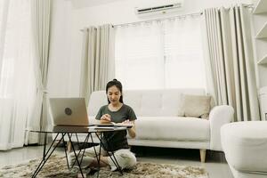 jonge aziatische vrouw zit in de woonkamer met behulp van een rekenmachine om gezinsinkomen en -uitgaven te berekenen en schrijft het thuis op in zijn notitieboekje of laptop. foto