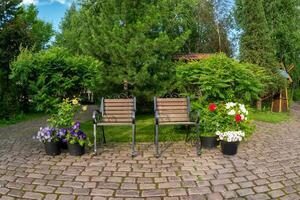 twee smeedijzer fauteuils in een zomer tuin met metselwerk van mooi bloeiend planten Aan een steen geplaveid voetstuk landschap ontwerp concept. foto