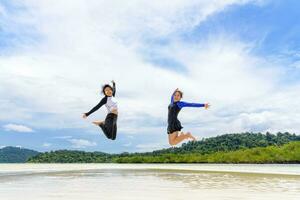 twee Aziatisch tiener meisjes vrienden jumping genieten Aan de strand foto