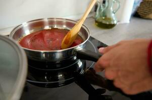 detailopname chef hand- gebruik makend van een houten lepel, kruiderij en menging een koken tomaat saus in een pan Aan elektrisch fornuis foto