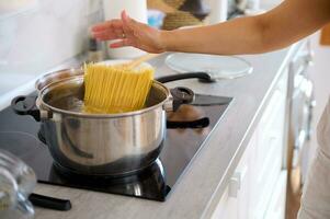 detailopname vrouw zetten spaghetti in een pan met een koken water, staand door een elektrisch fornuis in modern keuken. foto