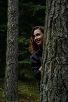 een jonge vrouw in een naaldbos kijkt uit achter een boom