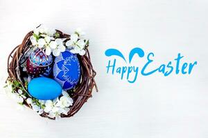 tekst gelukkig Pasen. nest met Pasen eieren en takken van voorjaar bloeiend kersen Aan een wit houten achtergrond. rogge eieren geschilderd met natuurlijk kleurstoffen. foto