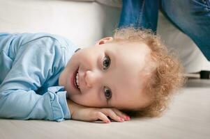 weinig meisje met gekruld haar- knipoogt. kind aan het liegen Aan een bank. portret van een jong glimlachen gelukkig baby meisje. schattig kleuter met versteld staan glimlachen gezicht foto