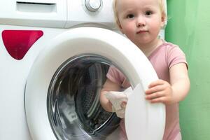 kleuter schoonmaak wassen puree. weinig meisje reinigt de Open deur van een wit het wassen machine Bij huis met vochtig lap. onderhouden netheid van huishouden huishoudelijke apparaten. foto