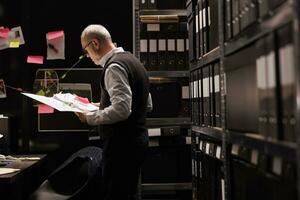 ouderen privaat detective werken Bij mysterieus crimineel geval, analyseren bestanden met tafereel misdrijf bewijs, controle slachtoffer rapport. Politie officier ontdekking potentieel verdachte in archiveren kamer foto