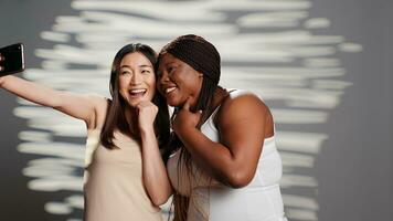 twee huidsverzorging modellen nemen afbeeldingen Aan smartphone, gebruik makend van mobiel telefoon naar hebben pret met foto's. jong mooi Dames bevorderen zelf liefde en lichaam positiviteit, zelf aanvaarding in studio. foto