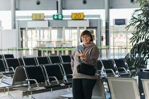 gelukkig glimlachen volwassen reiziger vrouw houdt smartphone, heeft video telefoongesprek, golven hand- in groet. oud vlogger of blogger heeft leven Bij luchthaven terminal aan het wachten voor vlucht, sociaal media. vakantie, vakantie foto