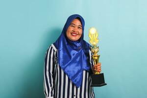 een opgewonden middelbare leeftijd Aziatisch zakenvrouw in een blauw hijab en een gestreept overhemd is glimlachen terwijl Holding een goud trofee, vieren haar succes en prestatie. ze is geïsoleerd Aan een blauw achtergrond foto