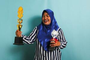 een opgewonden middelbare leeftijd Aziatisch zakenvrouw in een blauw hijab en een gestreept overhemd is Holding een goud en zilver trofee, vieren haar succes en prestatie. ze is geïsoleerd Aan een blauw achtergrond foto