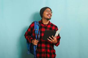 opgewonden jong Aziatisch leerling Mens met een muts en een rood plaid flanel shirt, vervelend een rugzak en bril, is op zoek terzijde Bij kopiëren ruimte terwijl Holding boeken en staand tegen een blauw achtergrond foto