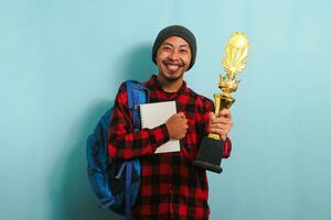 gelukkig jong Aziatisch Mens vervelend een rugzak, muts hoed, en rood plaid flanel shirt, Holding een goud trofee, verheugt in zijn succes en prestatie, geïsoleerd Aan een blauw achtergrond. foto