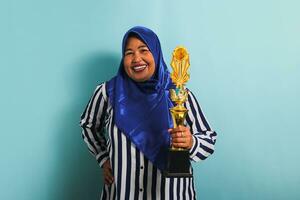 een opgewonden middelbare leeftijd Aziatisch zakenvrouw in een blauw hijab en een gestreept overhemd is Holding een goud trofee, vieren haar succes en prestatie. ze is geïsoleerd Aan een blauw achtergrond foto