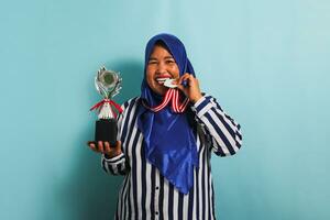 een gelukkig middelbare leeftijd Aziatisch zakenvrouw in een blauw hijab en een gestreept overhemd is bijten een medaille terwijl Holding een zilver trofee, vieren haar succes en prestatie, geïsoleerd Aan een blauw achtergrond foto