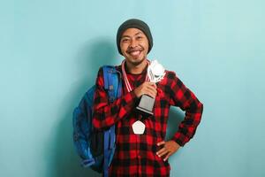 gelukkig jong Aziatisch Mens leerling tonen een medaille en trofee, geïsoleerd Aan een blauw achtergrond foto
