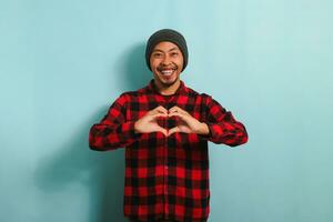glimlachen jong Aziatisch Mens shows een liefde gebaar met zijn hand- terwijl staand tegen een blauw achtergrond foto